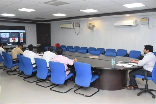 सम्मेलन कक्ष में मेज के चारों ओर बैठे लोग सामने लगी स्क्रीन पर वीडियो कॉन्फ्रेंस में भाग लेते हुए