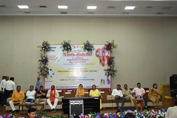 सभागार के मंच पर बैठे अतिथि, पीछे सहायक उपकरण वितरण शिविर का बैनर और फूलों की सजावट