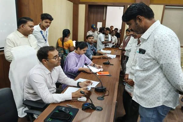 बैठक कक्ष में मेज के सामने खड़े व्यक्ति का आवेदन देखते अधिकारी और पास बैठी महिला अधिकारी